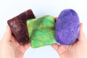 hands holding 3 felted soaps, one red with silk fibers, one different colors of green, and a purple round one